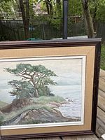 Close-up of coastal seascape painting showing detail of trees, rocky shoreline, and ocean water with signature visible.