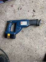 Blue 18 volt cordless reciprocating saw resting on concrete surface showing side profile including battery and blade attachment point.