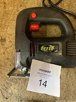 Black Tmt corded electric jigsaw with visible red trigger button and safety lock, clear plastic blade guard, power cord coiled beside it on a beige carpet.