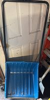 Blue plastic snow shovel with black metal handle leaning against a white garage door, some dirt visible in scoop.