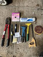 Overview of all tools and supplies laid out on concrete including bolt cutters, paintbrushes, wire stripper, scraper, handsaw, sanding discs, and wire roll