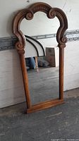 Full view of the large vintage mirror, showing the wood frame, carved ornamental top, and mirror glass reflecting the background.