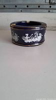 Side view of ceramic ashtray showing detailed classical scene silhouette in white on dark blue background with gold trim.