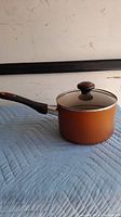 Orange Farberware non-stick saucepan with black handle sitting on a blue quilted surface, showing side profile and spout.