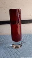 Tall cylindrical red glass vase with thick solid base, photographed on blue padding and against a white background.