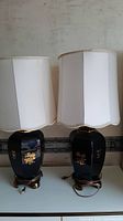 Two black ceramic table lamps with gold floral and calligraphy patterns, white scalloped fabric shades, and brass metal bases, shown side by side.