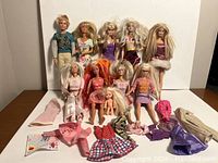 Photo shows 10 dolls including 9 Barbie dolls and 1 Ken doll along with multiple clothing pieces laid out on a table.