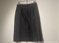 Photo of black textured women's skirt hanging on a hanger against a plain wall.