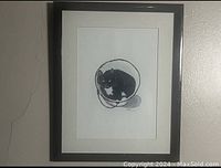 Framed black and white picture of a cat inside a round container, showing the entire framed item hung on a wall.