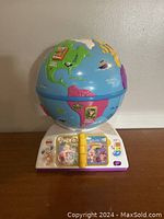 Front view of a colorful Fisher Price Laugh & Learn globe showing North and South America with various animals and landmarks. The globe sits on a white base with purple trim featuring interactive components including buttons and a small flip book in yellow.