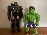 Photo showing Iron Man black and silver armored superhero figure and green Hulk action figure standing side by side on a flat surface.