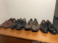 Five pairs of men's shoes size 12 lined up on a surface against a wall showing front and side angles. Includes tassel loafers, black dress shoes, suede lace-ups, and brogue shoes.