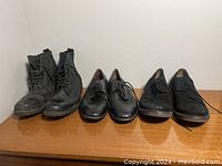 Four men's shoes size 12.5 displayed on a wooden surface against a white wall, including black lace-up boots, black leather dress shoes, and black suede shoes.