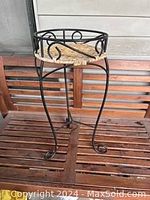 First image showing full view of one wicker and metal plant stand on a wooden slat surface against a wooden paneled background.