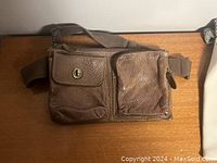 Brown leather crossbody bag with two front pockets and adjustable fabric strap lying flat on wooden surface.