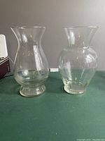Two clear glass vases shown standing side by side on a green surface with a plain background, front view