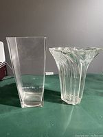 Side view of two clear glass vases on a green surface, one tall square vase and one wavy rim vase with ivy pattern