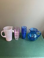 Wide shot showing pink pitcher, four pink tumblers, blue pitcher, and four unopened blue tall glasses on table.