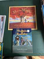 Front cover of 'M is for Maple' and 'The Pirates of Georgian Bay' softcover books lying side by side on a green surface.