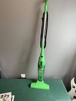 Full view of assembled upright Bissell vacuum cleaner in green with black power cord wound around handle, standing on green table against beige wall.