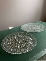 Two clear glass plates with bubble textured bases placed side by side on a green surface. One plate is larger (14 inch) and the other smaller (12 inch). Light reflection visible on textured bubbles.