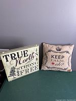 Photo showing the front of a wooden wall hanging with text 'True North and Strong Free' and a 15x15 faux suede decorative pillow with 'Keep Calm, eh?' text.