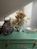 Wide view showing brown metal lantern, blue vase with faux flowers, and glass bowl on gold metal stand on green chest