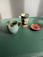 Photo of all three pottery items on a green surface: small grey jug, handpainted cup with person design, bright floral plate.