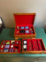 Open wooden jewellery box showing three interior trays lined with red velvet containing assorted jewellery items including necklaces, earrings, bracelets, and rings.
