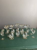 Full view of 17 glasses arranged on a green cloth: 5 champagne glasses, 6 sherry glasses, 6 brandy glasses all with gold trim.