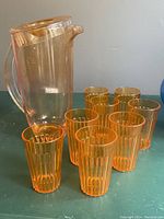 Orange plastic pitcher with lid, placed behind 8 small orange plastic glasses with vertical ribbing, all arranged on green surface.