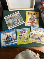 Photo showing the five paperback children’s storybooks arranged on a green surface: 'Where the Wild Things Are', 'Braids!', 'Fly Away Home', and two 'Why Should I?' books titled 'Protect Nature' and 'Recycle'.