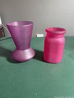 Two vases side by side on green surface; purple vase taller and tapered, pink vase shorter and cylindrical.
