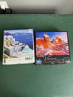 Two unopened boxes of puzzles with artwork shown: 550-piece Santorini scene and 1000-piece red rock formations.