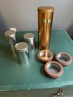 Lot of seven round metal tins including three silver tea tins, one tall gold pasta tin, and three smaller screw-top tins with clear window lids on a green surface.