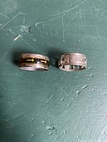 Top view of two silver-tone rings on green surface: one with yellow and black arrow engravings, the other with text engraving and cutouts.