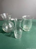 Three clear glass vases on a green surface against a gray wall, showing various shapes and sizes