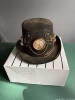 Front view of steampunk top hat showing clock face and metal cogwheel decorations on black fabric.