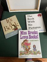 Three children's books about books displayed together: 'It's a Book' by Lane Smith, 'The Book With No Pictures' by B.J. Novak, and 'Miss Brooks Loves Books (and I Don't)' by Barbara Bottner with illustrated cover.