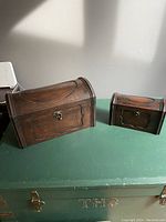 Both wooden chests placed side by side on a green table surface, showing size difference and front panel design.