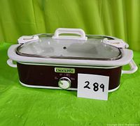 Front view of the Crock-Pot slow cooker showing brown body, white handles, clear glass lid with white handle, control knob, and label.