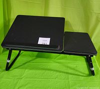 Foldable black laptop tray desk with two surfaces and adjustable legs, shown from front angle on green background.