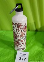 Photo of the white metal water bottle with red and brown floral design, black lid, and attached silver carabiner on green background.