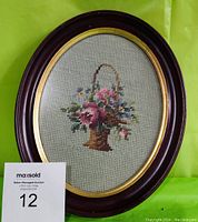 Front view of the handmade needlepoint floral basket framed under glass in an oval wooden frame with dark finish and gold trim.