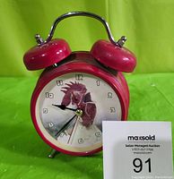 Front view of red twin bell alarm clock showing rooster image on clock face and clock hands set to about 8:40:37.