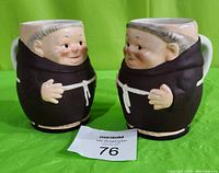 Front view of both vintage Hummel Goebel monk mugs side by side showing detailed faces and bodies with the monk robes and white rope belt, placed on green background with auction tag in front.