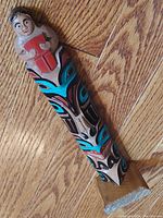 Close-up angled view of the carved and painted human figure holding a red item at the totem pole's top, mounted on a wooden base on hardwood floor.