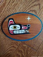 Photo of oval wooden plaque on a wooden floor, showing the painted stylized wolf head with star symbol.