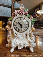 Frontal view of the porcelain cherub clock showing clock face with Roman numerals and cherubs on each side along with floral details on top.