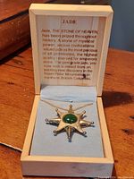 Close-up of the necklace pendant with green jade center on gold-tone starburst setting inside blue velvet jewelry box with printed description inside lid.
